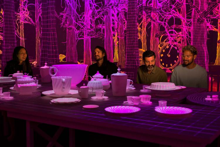 People sit at a giant table set for afternoon tea against an illuminated backdrop of pink and purple trees
