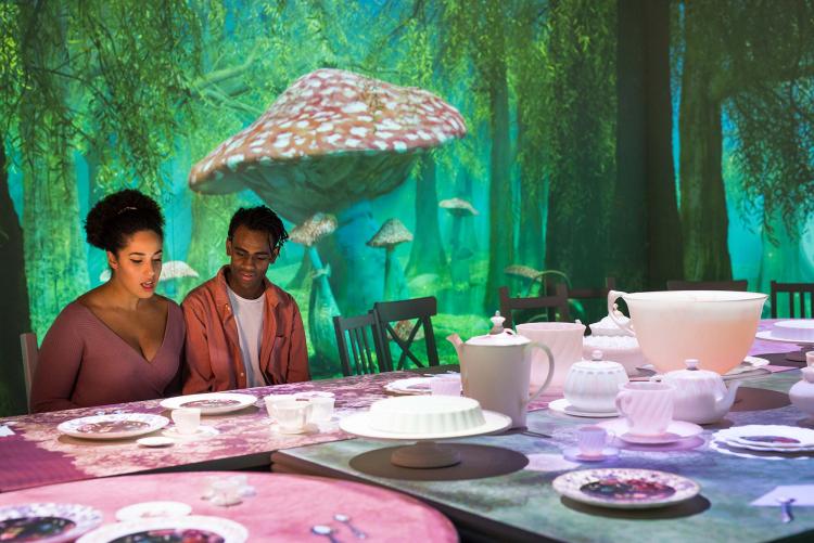 Two people sit at a giant table set for afternoon tea against an illuminated backdrop of greenery and mushrooms