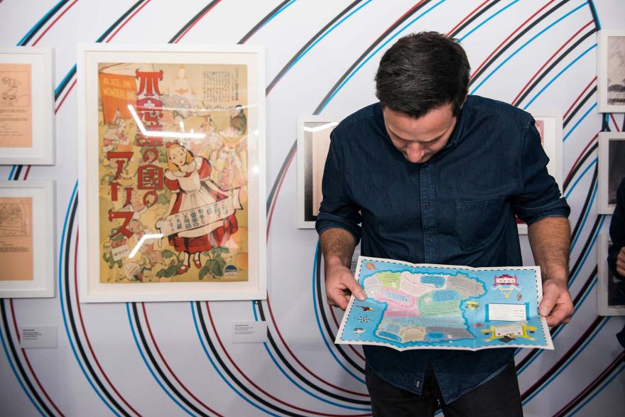 A man reading a map leans against a wall decorated with hypnotic spiral patterns and illustrations from Alice in Wonderland