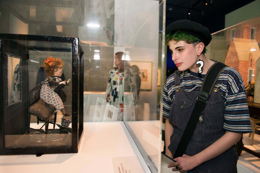 A person wearing large question mark earrings examines a doll in a museum showcase