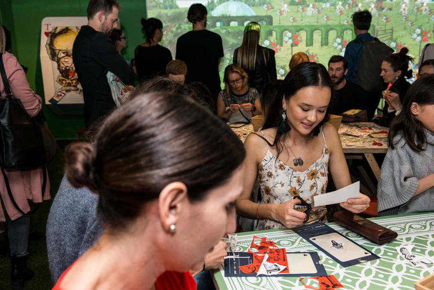 People sit at long tables decorating large playing cards, in front of an illuminated backdrop depicting the Queen of Hearts' garden from Alice in Wonderland