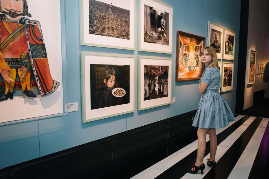 A blonde girl wearing a blue dress stands in front of a display of Alice in Wonderland artworks