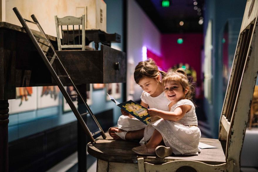 Two children sit on an enormous chair in front of an equally large desk, reading a map of Wonderland