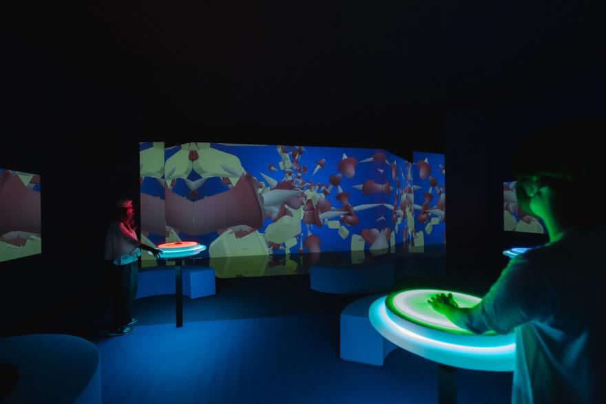 People stand at glowing podiums surrounded by screens filled with kaleidoscopic images