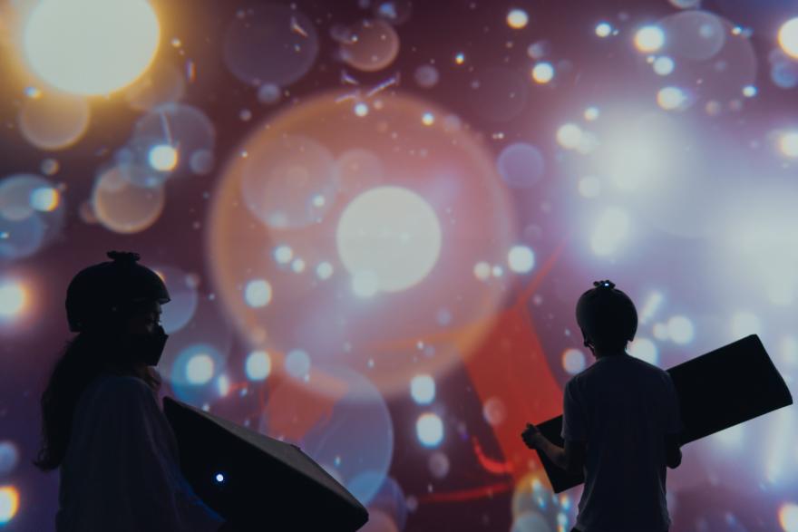 People holding large geometric shapes gaze look up at projected points of light all around them