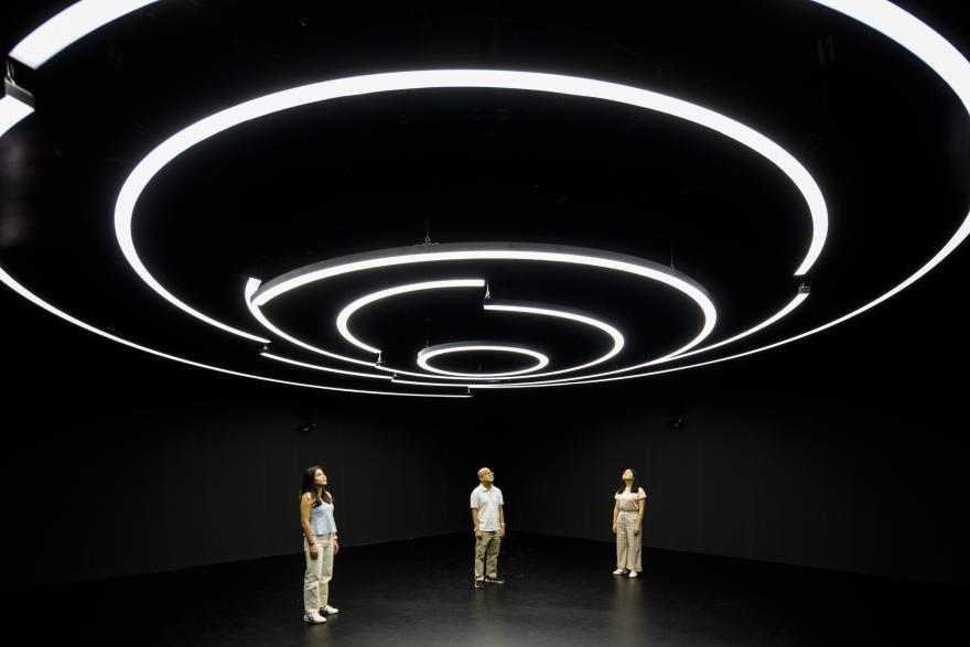 People standing in a large dark space gaze up at glowing white concentric circles on the ceiling