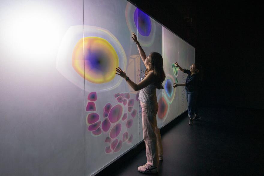 People touch floating amoeba-like projections on a glowing white wall