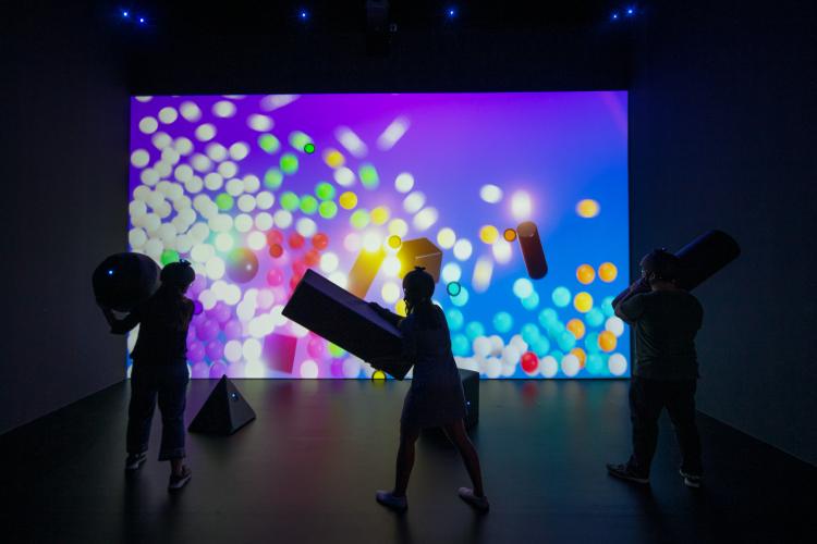 Three people wearing helmets are facing a giant colourful wall filled with shapes. They hold giant soft toy shapes, moving them to interact with the screen. 