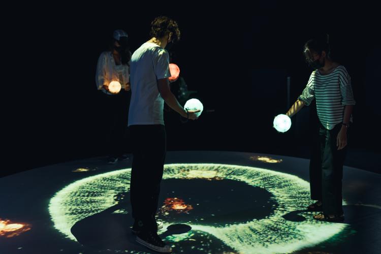 Three people hold coloured orbs, looking down and moving with a circular projection of light on the floor. 