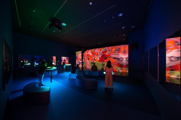 Three people spin illuminated neon freestanding discs, changing the colourful projections on a screen opposite them, while three people sit to watch the projections unfold.