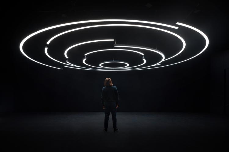 A man stands alone in the middle of a dark room, staring upward at circles of light caused by neon bars overhead.
