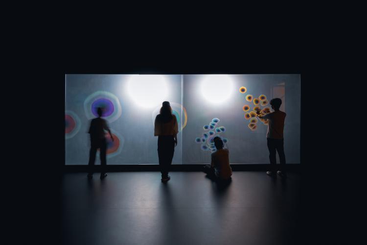 Four people stand evenly spaced in a row, facing a screen lit with colourful circles against a white backdrop, the circles responding to their unique movement. 