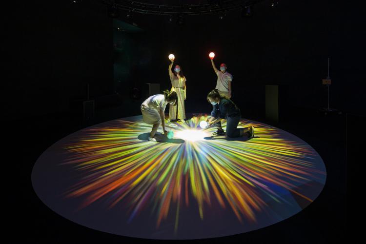  Four people stand together at various levels, holding lit orbs above their head while standing on a burst of star-like colour on the floor. 