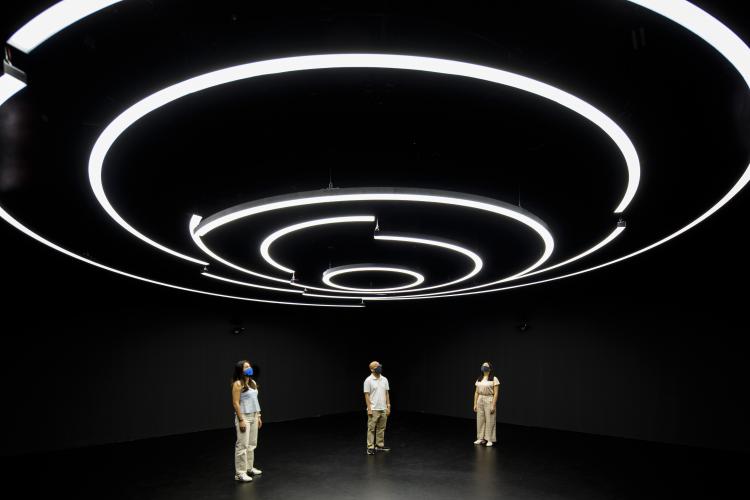 Three people wearing facemasks stand in the middle of a dark room, staring upward at circles of light caused by neon bars overhead.