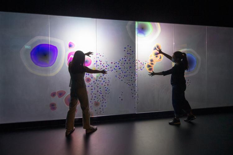  Two women lean towards each other, facing a screen lit with colourful circles against a white backdrop, the circles responding to their unique movement. 