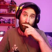 Wayward Sinny, a thirty something bearded man, wears a headset and leans on his hand at a computer desk, looking into the camera