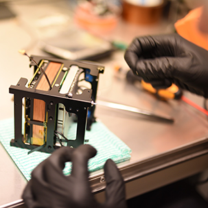 A pair of black gloved hands working on a piece of technology in the shape of a cube