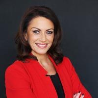 Rabia Saddique, a middle eastern woman wearing a red blazer with her arms crossed, smiles warmly at the camera