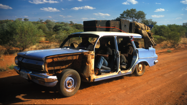 An old blue car with luggage on the top and four passengers drives over red dirt with blue skies overhead in the Australian outback.