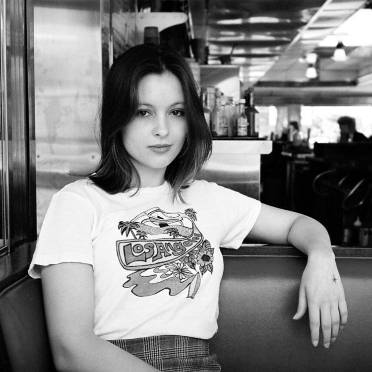 Black and white image of a person with dark brown hair, white t-shirt with band logo and jeans resting on a seat with a cafe setting behind her.