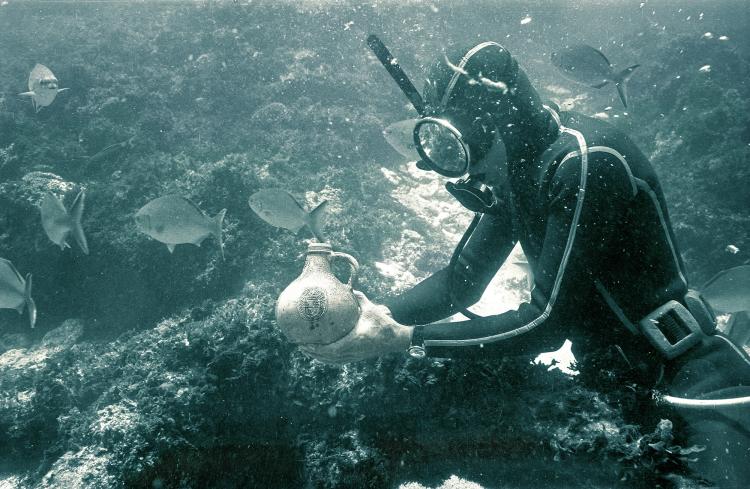 A diver underwater at a shipwreck site examines a pottery jug