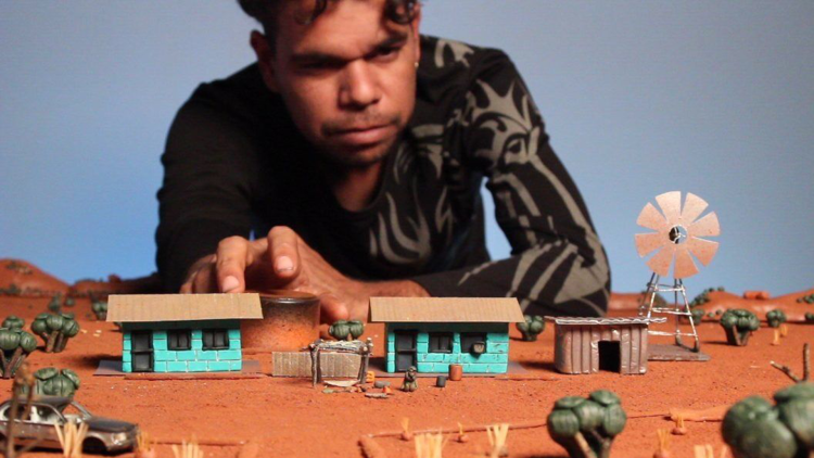 Jason wears a black patterned long sleeve shirt and leans over a miniture model of an outback town made from paper and clay he created.