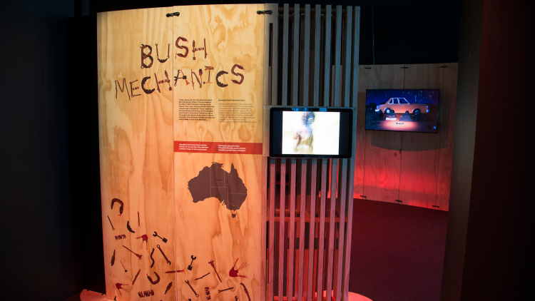 A wood panel stands in the gallery entrance with the Bush Mechanics title printed on it, a small TV screen is fixed to the panel on the right hand side with an additional screen seen in the distance behind.