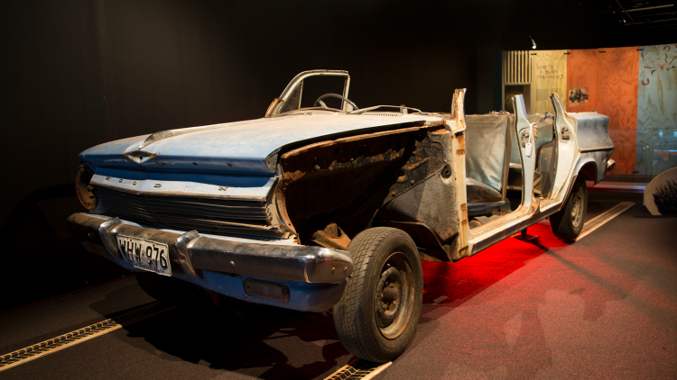 An old blue car sits on display with the roof missing and the left handside removed revealing the interior.