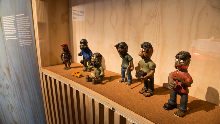 Six clay figures wearing different coloured pants and tops perch on a wooden surface on display, each in different positions depicting character moments from the TV series.