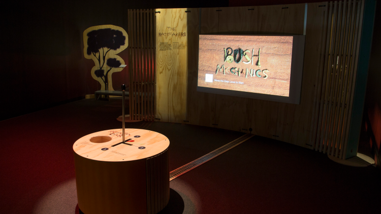 A circular wooden table sits centrally with interactive parts built into it, facing a TV screen with rthew Bush Mechanics logo displayed, a wooden tree cut-out to the left hand side of the screen.