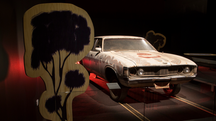 A white Ford Fairlane car painted with traditional Warlpiri designs in orange and silver is centrally lit in a dark space, with a wooden tree cut-out in the foreground.