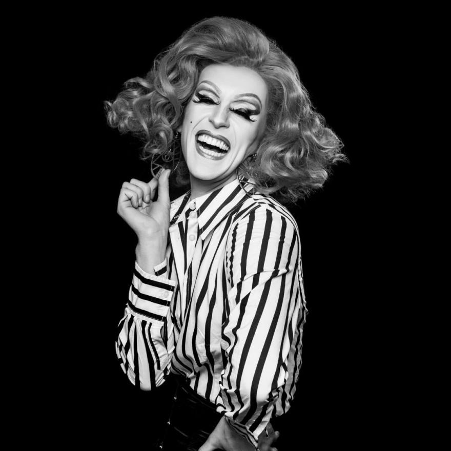 Black and white image of a Drag Queen dressed in a stripey white and black shirt and big curly hair.