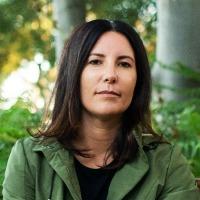 Elfie Shiosaki wears a green jacket over black clothing with dark, straight medium length hair. She sits on a wooden bench facing forwards, surrounded by green rainforest.