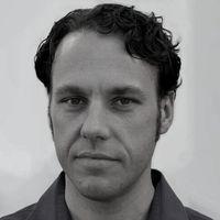 Black and white headshot of a man with dark hair wearing a black shirt facing the camera with a white wall in the background