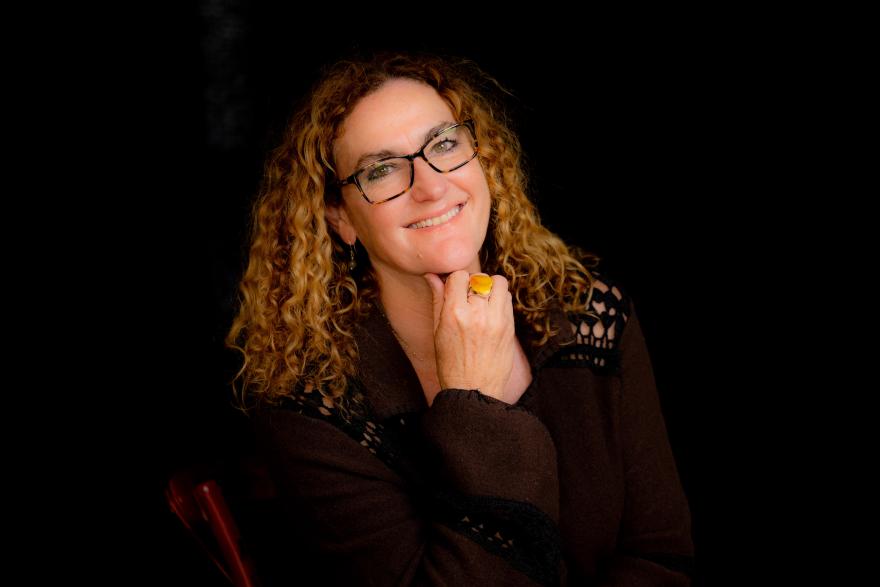 Woman with shoulder length hair and glasses smiles and looks happy with right hand close to face 