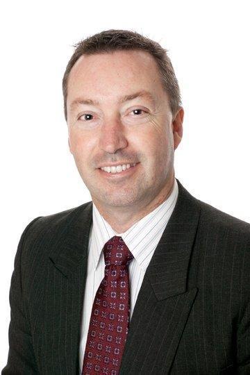 Man with short dark hair, white collar shirt, dark red tie and blazer smiles