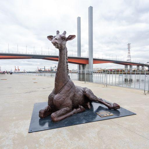 A large bronze sculpture of a baby giraffe sitting down in the city