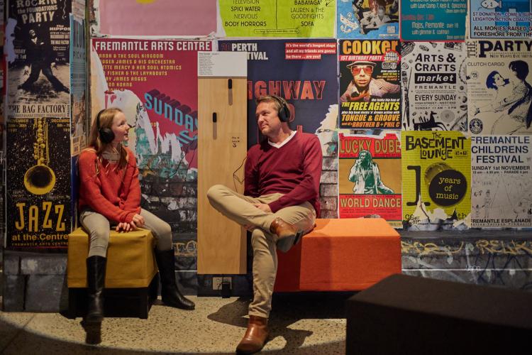 Image of an older man and younger girl sitting on stools and listening to exhibition audio through headphones, surrounded by colourful posters of varied Fremantle arts events