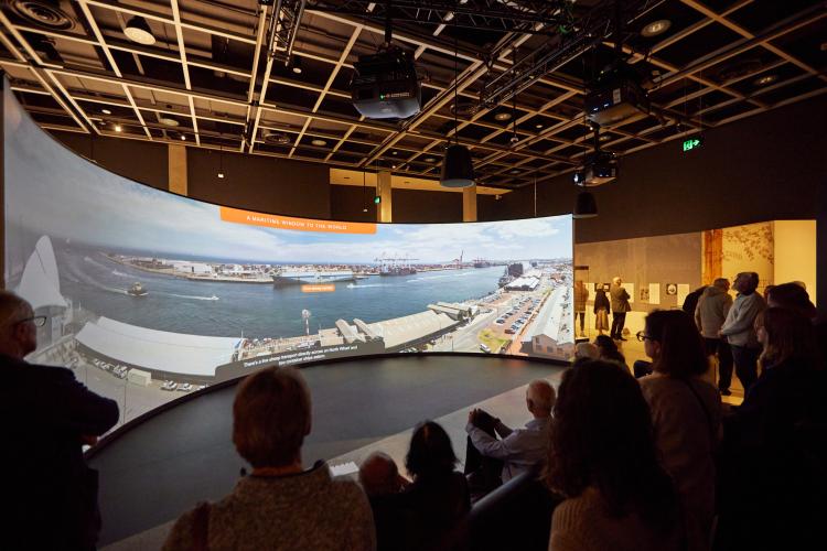 An image of a small crowd gathered around a large curved projection screen which displays a panoramic view of Fremantle's maritime port with the title reading A Maritime Window to the World