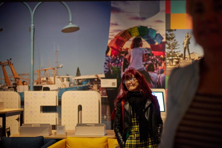 An image of an older woman in a yellow tartan dress andf leather jacket standing in front of a wall of colourful images of Fremantle including a scultpure of shipping contained arranged as a rainbow and a large ship. 