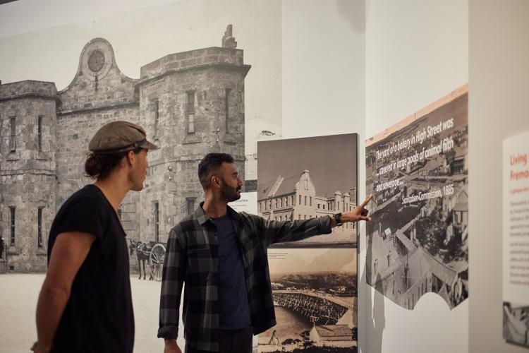 An image of two men in a photo gallery surrounded by black and white images of Fremantle buildings including the Fremantle Prison gatehouse and Fremantle Arts Centre. One man is pointing at a quotation on the wall while another man watches on. 