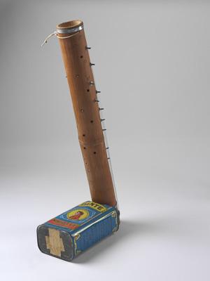 A hybrid instrument stands upright, composed of a tin can on the bottom, and a long brown circular pipe protruding upwards. Attached to the pipe are a series of strings and dials.