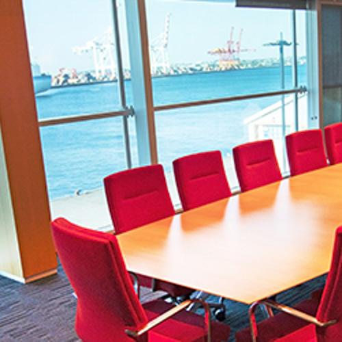 Red office chairs are positioned around a light brown table in a boardroom-style set up. Through the large brightly-lit window behind the set-up is an expansive view of Fremantle harbour.