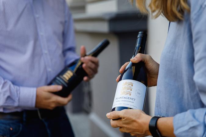 An image taken of the torsos of two people, both are wearing blue button-up skirts and holding bottles of wine. The bottle in the foreground displays a white label with three decorative lions and the words Plantagenet York Chardonnay