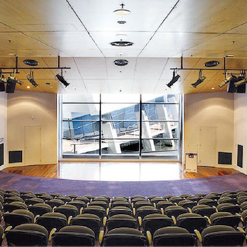 An image taken from the back of the Shipping Theatre overlooking rows of black chairs, angled towards a large square window through which a large white building is visible.