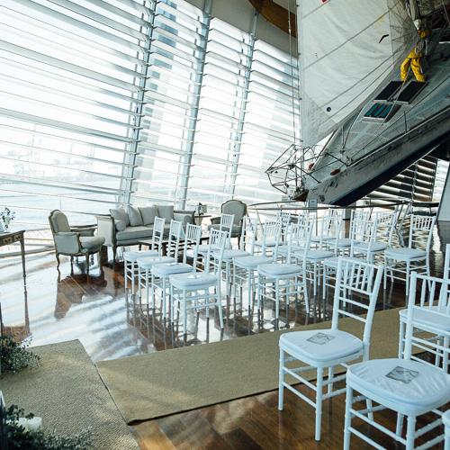 A wedding ceremony set up in the Australia 2 gallery at the Maritime Museum featuring white chairs in rows along an aisle, with arm chairs around the edge of the area and the bow of the Australia 2 ship visible in the background