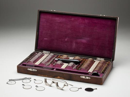 A case lined with a red-purple velvet-like material lies open, displaying a box of trial optical lenses and frames in individual slots in the case. 