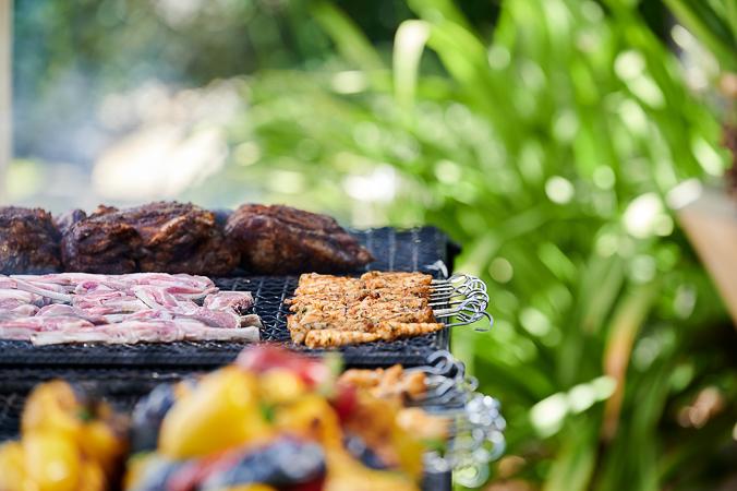 An open grill with colourful vegetable skewers and a variety of meats stands in focus behind a blurred garden background