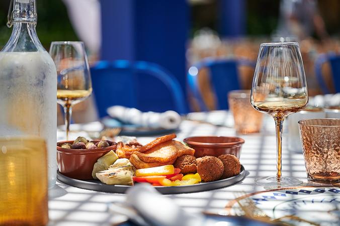 A checkered table cloth with several amber-tinted wine and water glasses with a platter of mediterranean foods including colourful vegetables, olives and kofta balls.