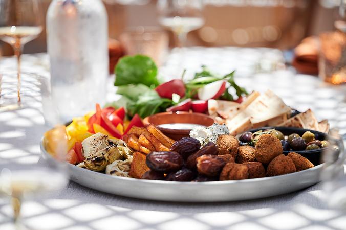 A checkered table cloth with several amber-tinted wine and water glasses with a platter of mediterranean foods including colourful vegetables, olives and kofta balls.